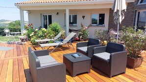 Un patio con mobiliario de mimbre, una mesa y sillas, rodeado de plantas en macetas y una casa con techo de tejas.