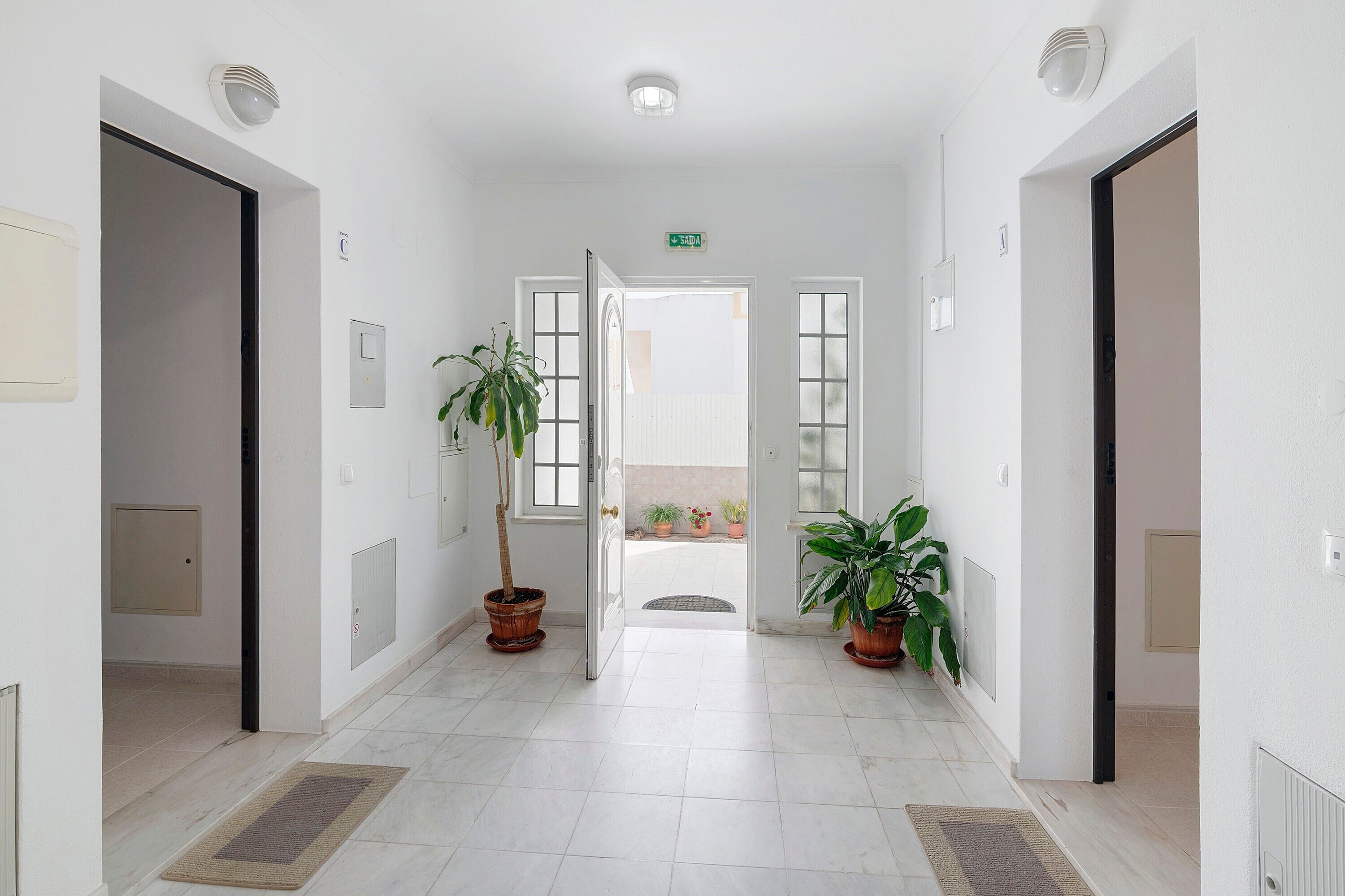Un couloir aux murs blancs, doté d’un sol carrelé et agrémenté de plantes en pot.