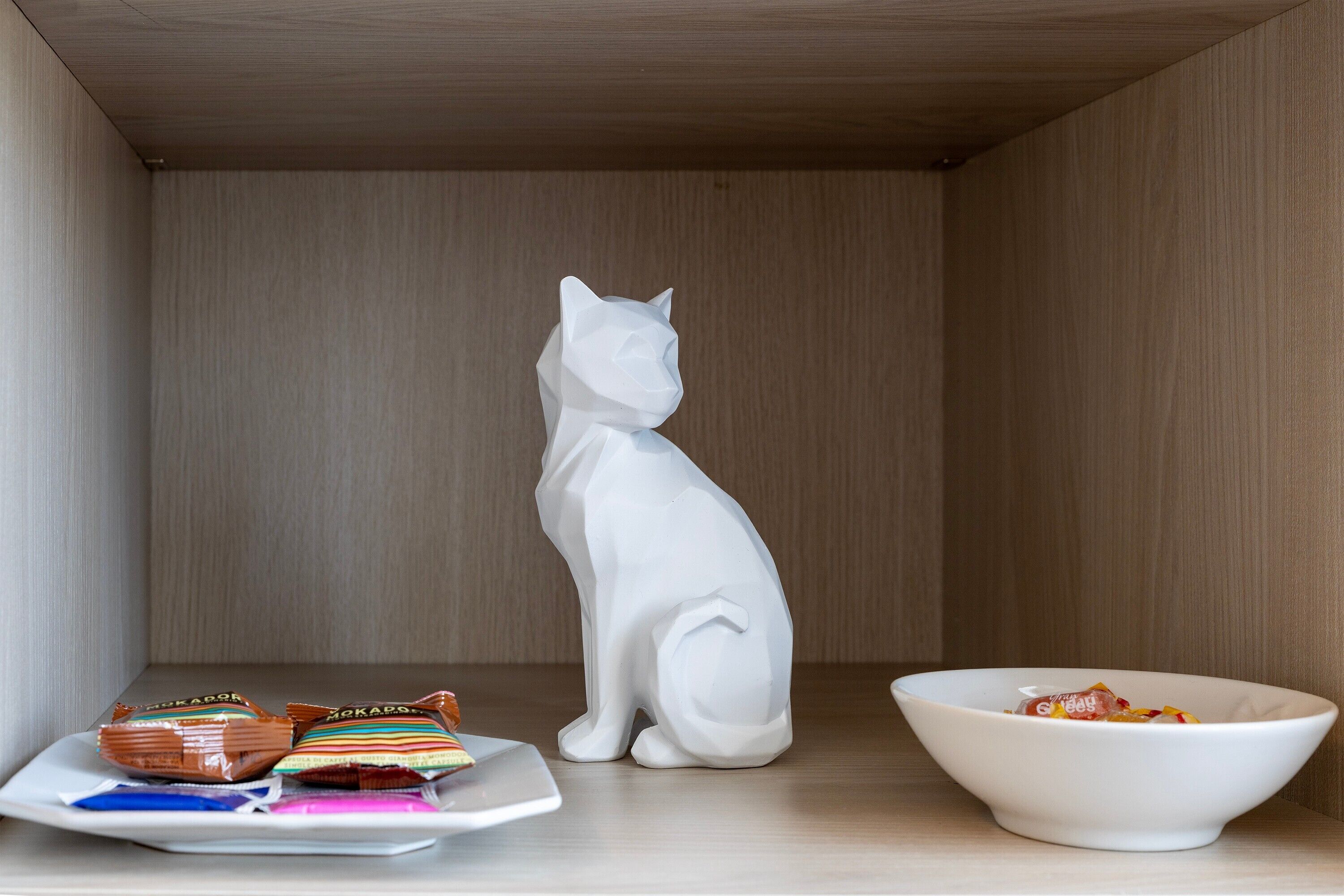 Een witte keramieken kattenbeeldje op een houten tafel met een schaal en porties snacks.