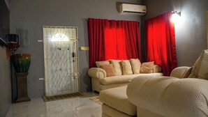Una sala de estar con un sofá color beige, cortinas rojas, un ventanal de vidrio y un equipo de aire acondicionado.