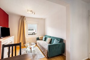 A compact room with a sofa, a TV on a stand, a dining table, and a window with a view of a car outside.