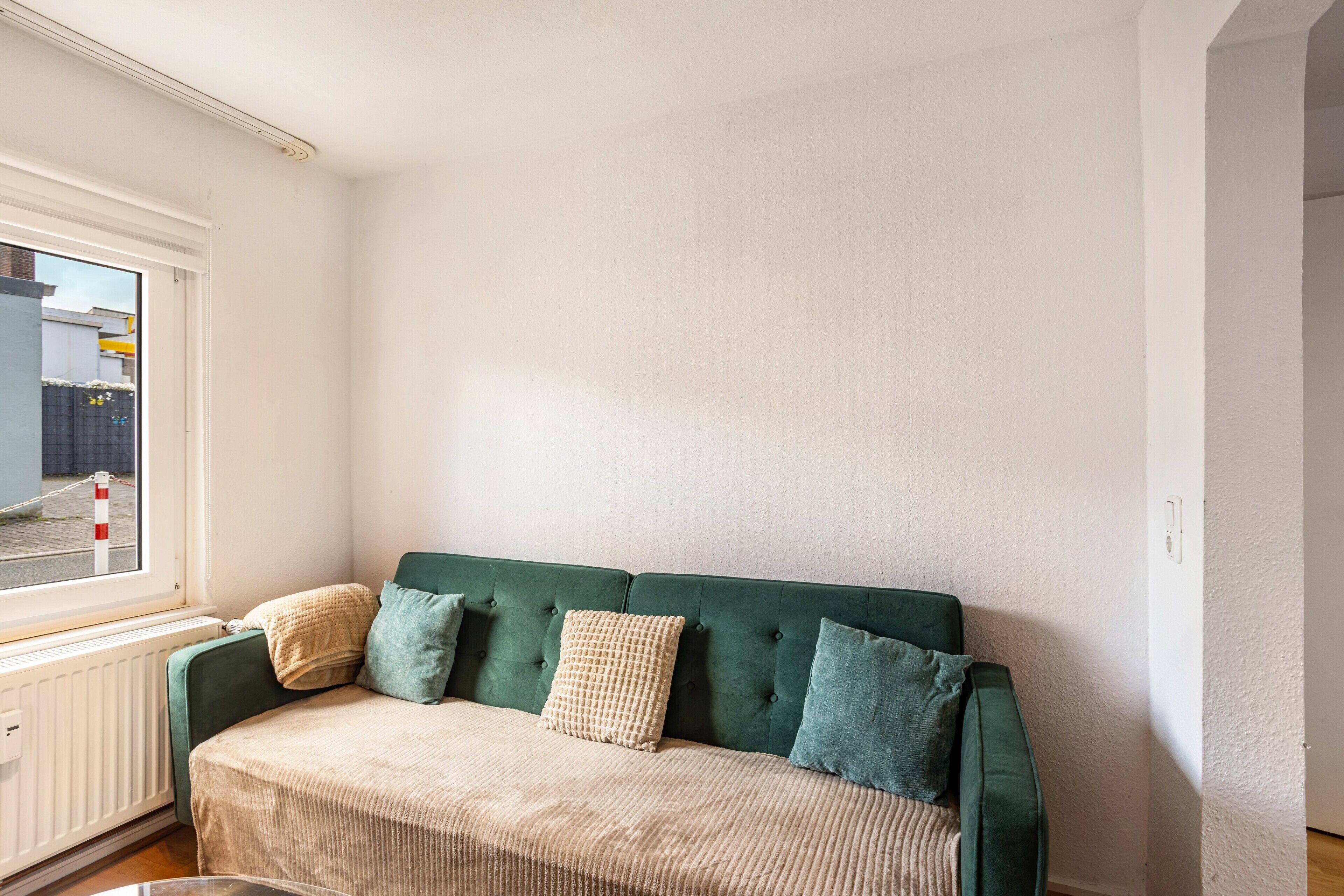 Un canapé vert avec un coussin beige, une fenêtre donnant sur une rue et un mur blanc.