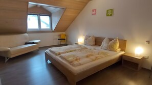 A bedroom with a sloped ceiling, a bed with patterned bedding, a sofa, a chair, and two wall-mounted lights.