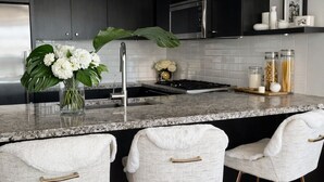 A modern kitchen with dark cabinets, a marble countertop, a gas stove, and three white bar stools with gold handles. - Ocean View Downtown (White Rock)