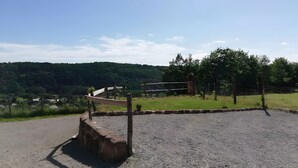 Een parkeerplaats met grind, omheind door een houten hek en poort, met veel groen en een glooiend landschap.