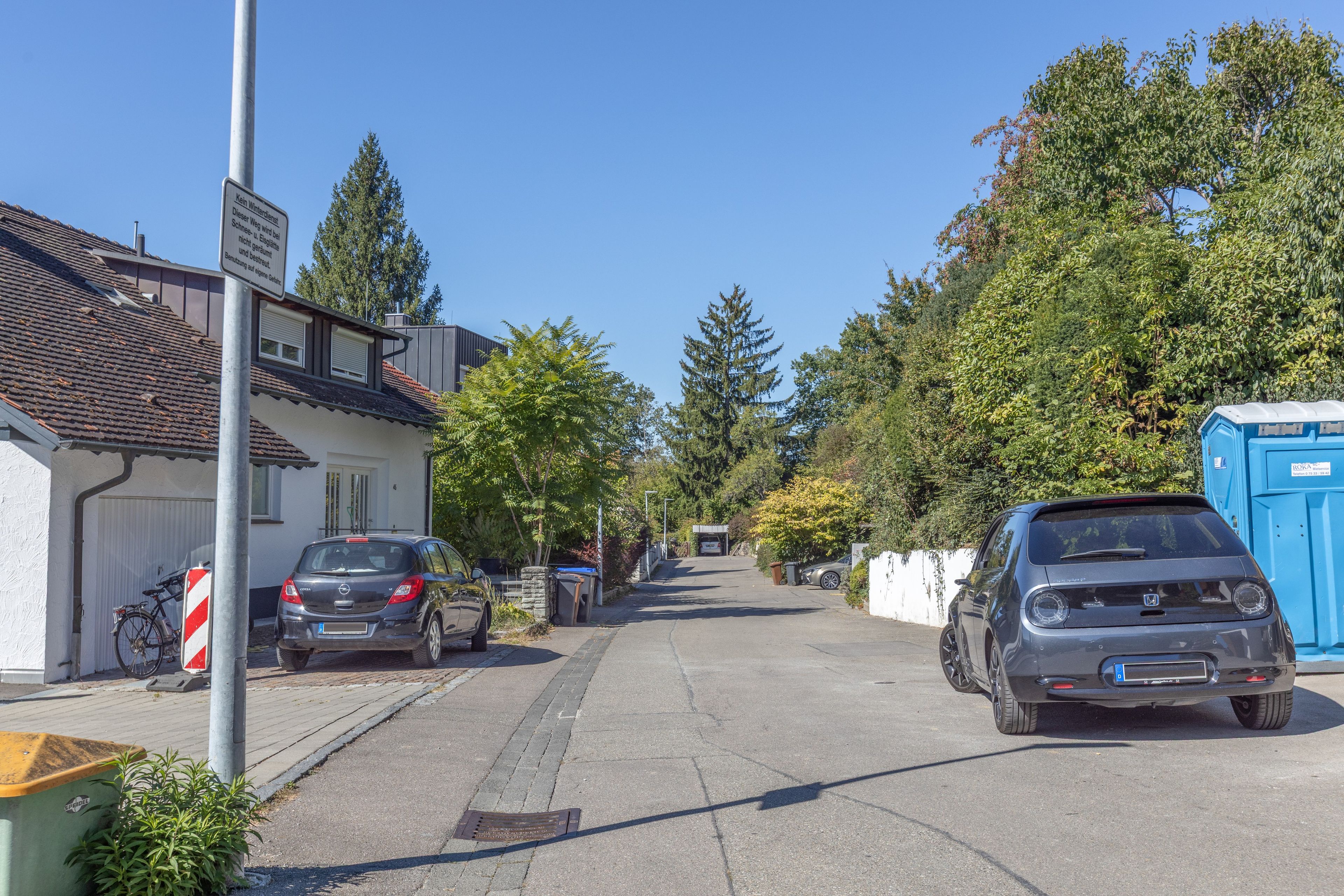 Eine Wohnstraße mit geparkten Autos, einer blauen mobilen Toilette und einem Fahrrad.
