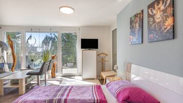 Une chambre d’hôtel moderne équipée d’un lit, d’un bureau, d’une chaise, d’une télévision et d’un balcon offrant une vue sur la verdure.