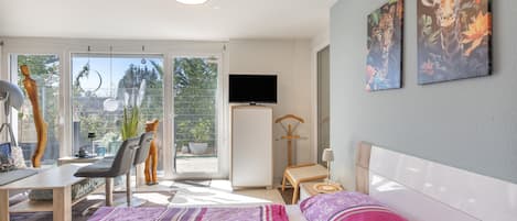 A modern hotel room with a bed, a desk, a chair, a TV, and a balcony with a view of greenery.