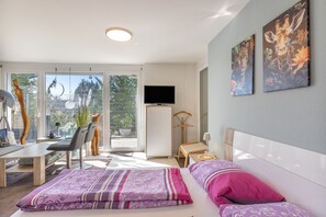 Een moderne hotelkamer met een bed, een bureau, een stoel, een televisie en een balkon met uitzicht op groen.