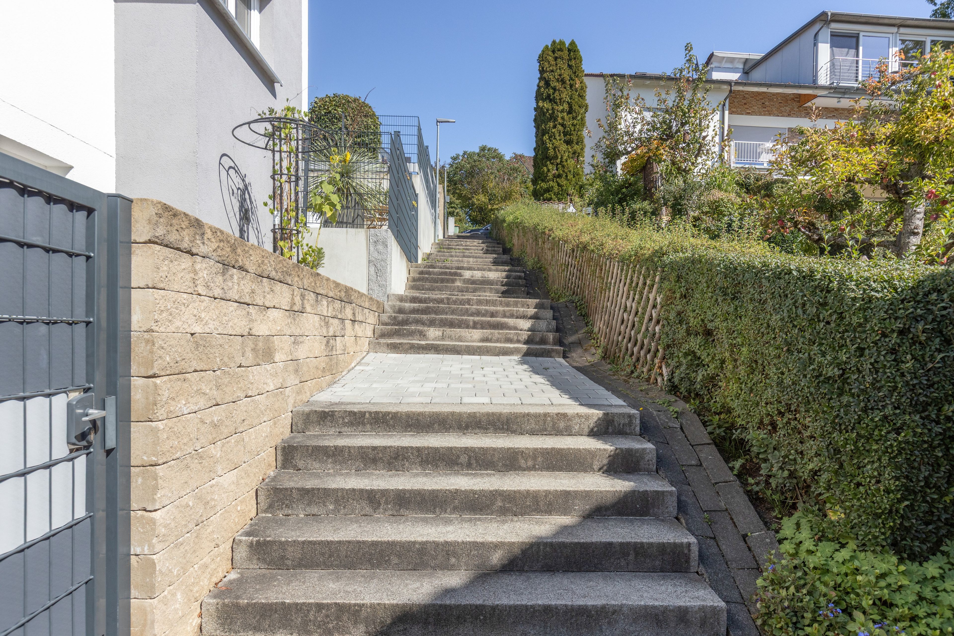 Eine Treppe aus Stein führt zu einem verschlossenen Eingang, begrenzt von einer Hecke und einem Gebäude mit weißer Fassade.