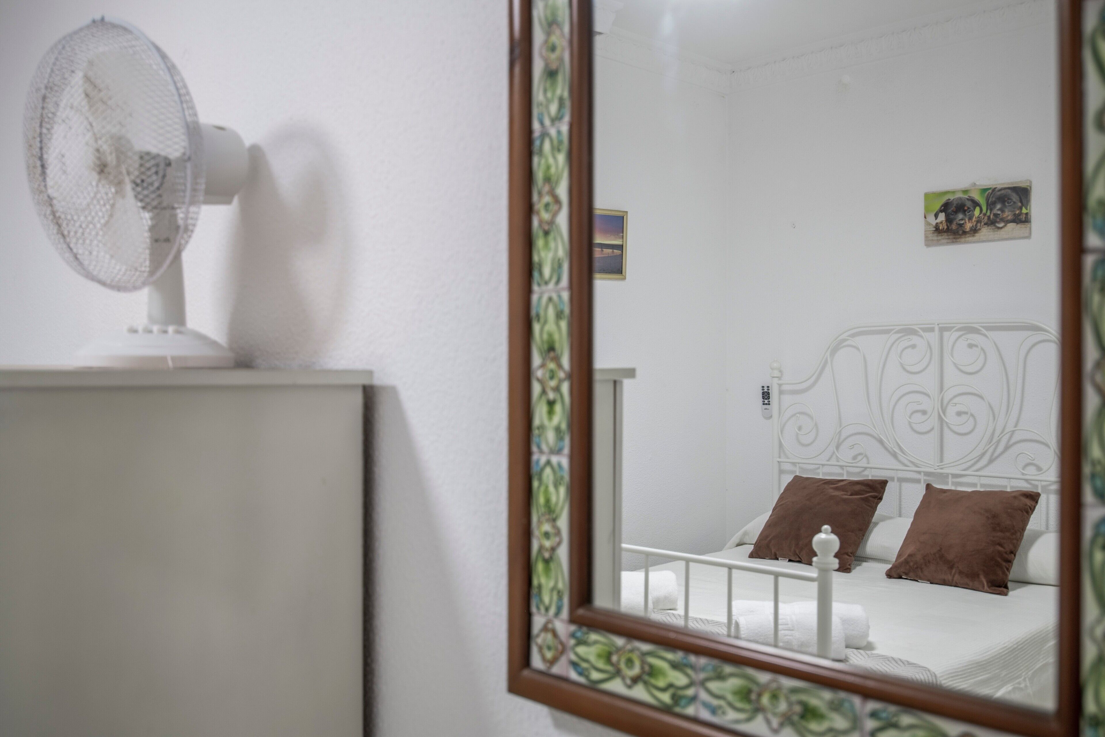 A bedroom with a bed, a white fan, and a mirror reflecting the room.