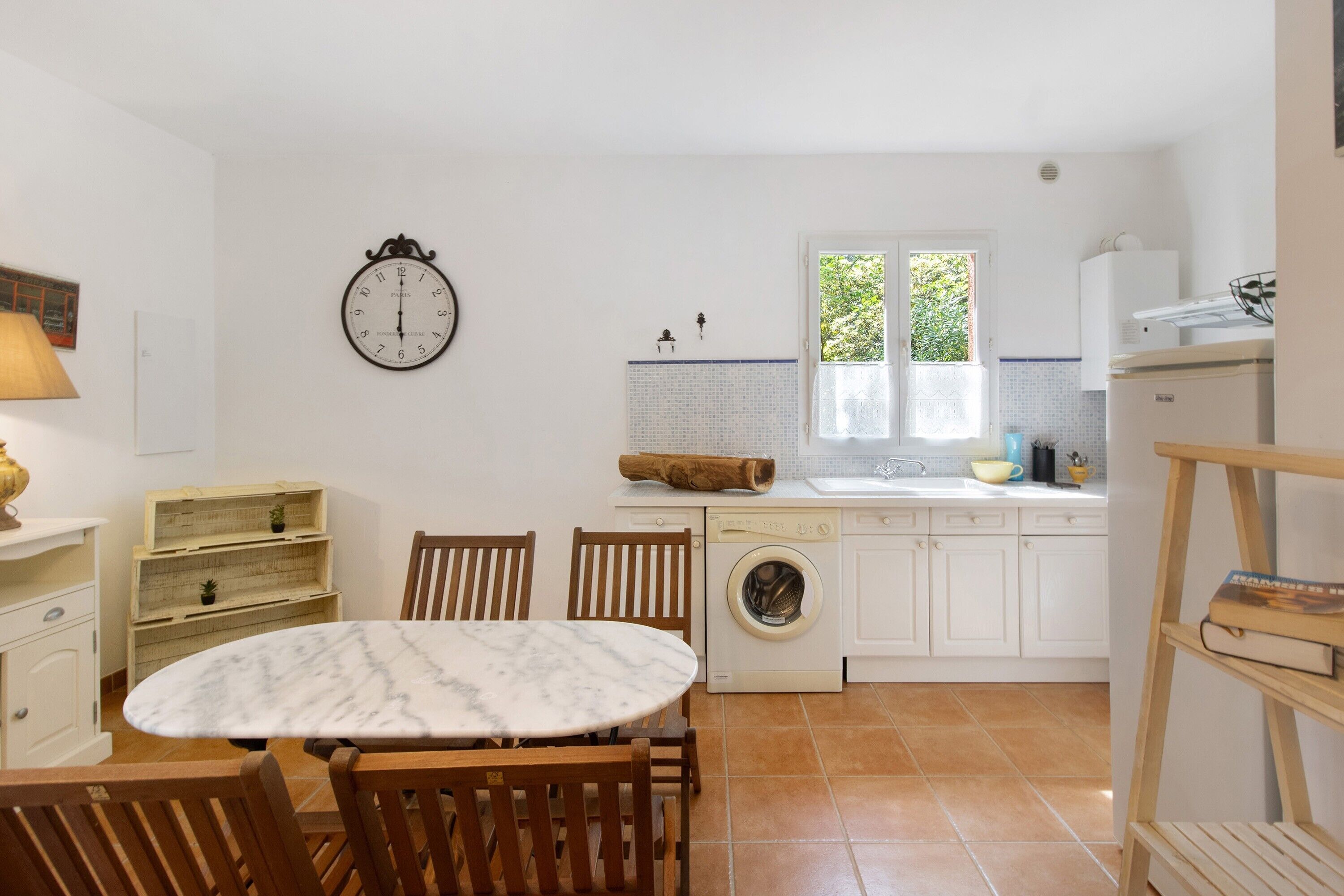 A compact kitchen with a washing machine, a dining table with chairs, and a clock on the wall.