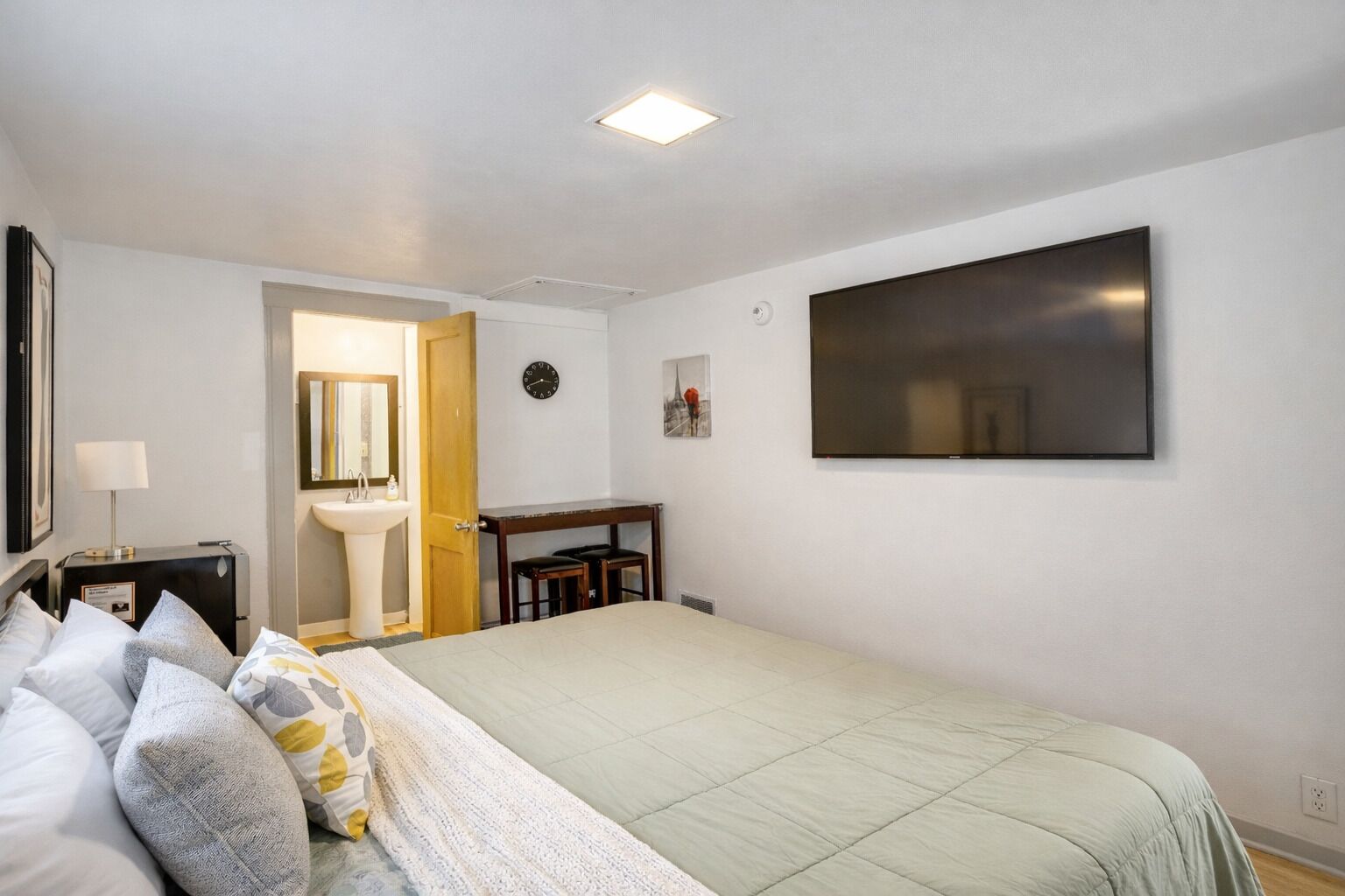 A bedroom with a bed, a bedside table, a nightstand, a wall-mounted TV, a clock, and a door leading to a bathroom.