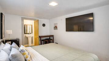 A bedroom with a bed, a bedside table, a nightstand, a wall-mounted TV, a clock, and a door leading to a bathroom.