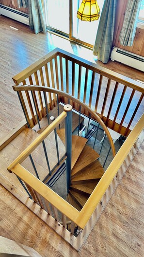 Una escala espirala de fusta amb baranes de vidre en una habitació amb sòl de fusta i una finestra amb cortines.