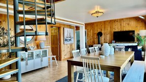 Ein modernes Interieur mit einer Holztreppe, einem Essbereich mit weißen Stühlen, einem Fernseher und einem großen Fenster.