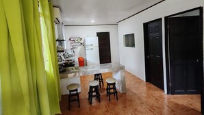 A kitchen with a white refrigerator, a sink, and a countertop. There are three stools and a small table. The room has a tiled floor and a door on the right. - Howler Monkey Sunset House / Just 200 mts from the Beach and Sunsets (Guanacaste Province)