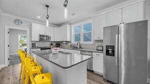 A modern kitchen with white cabinets, a stainless steel refrigerator, a large island with bar stools, and a window with blinds. - Golf Cart Included + Close to Beach and Restaurants! (Carolina Beach)