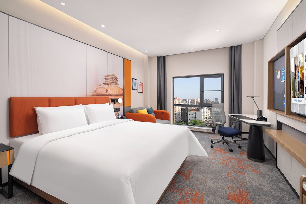 Une chambre d’hôtel avec un grand lit, un bureau, une chaise, une vue sur la ville et une grande fenêtre.