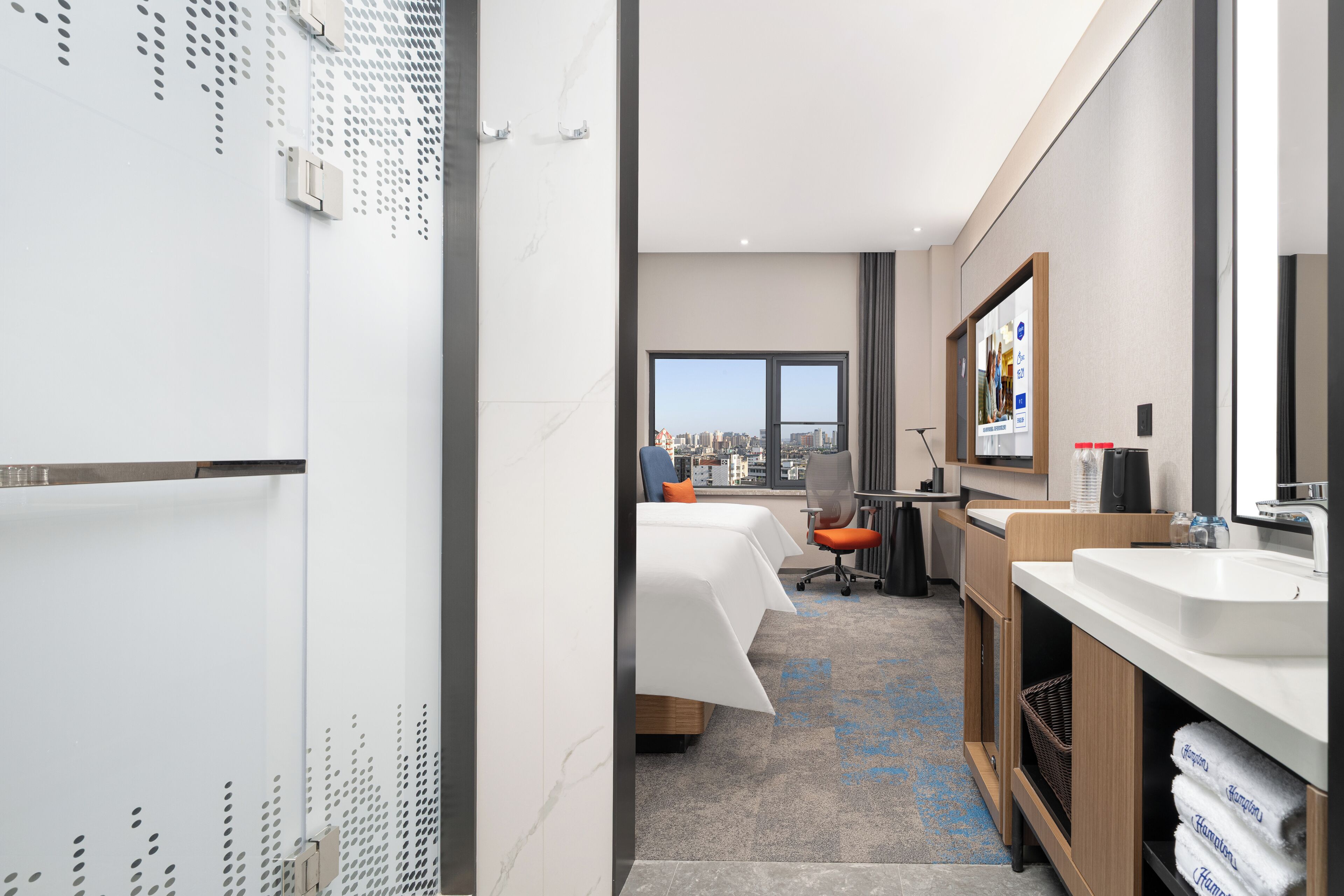 A hotel room with a bed, desk, chair, and a large window offering a city view.