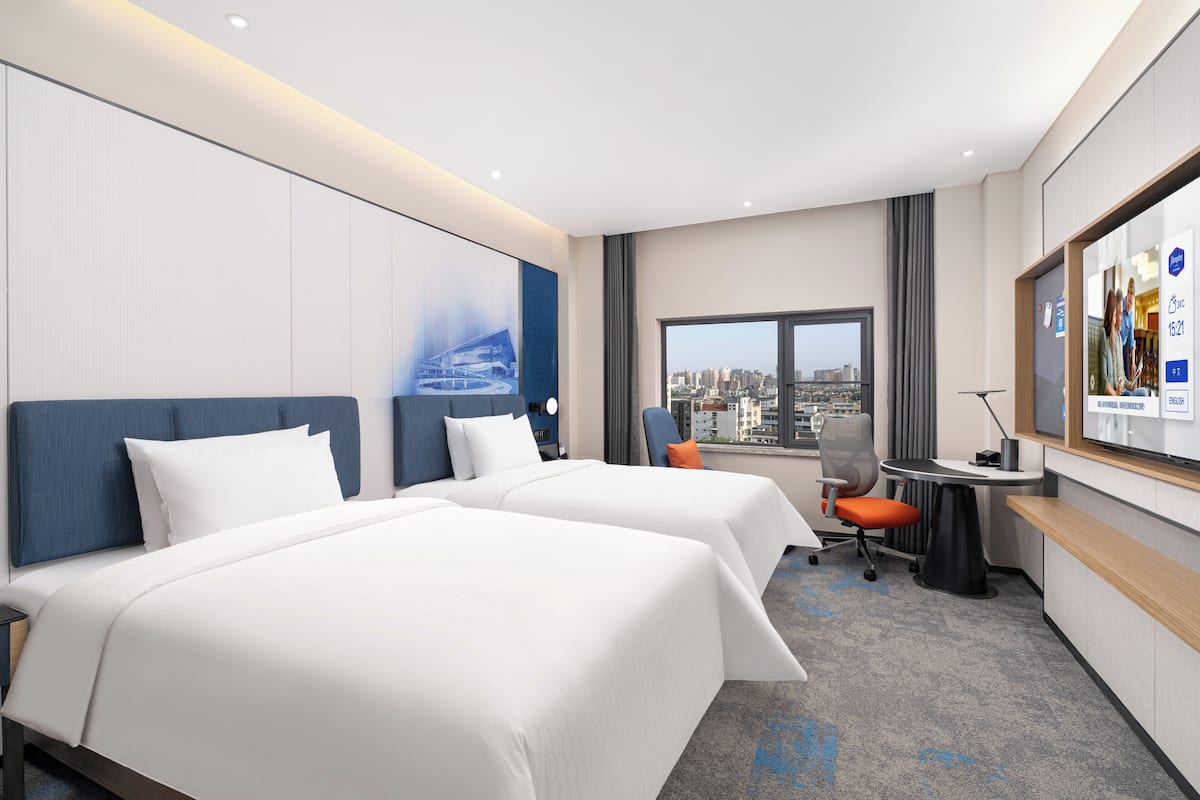 Une chambre d’hôtel avec deux lits, un bureau, une chaise, une grande fenêtre donnant sur la ville et un écran de télévision affichant des informations.