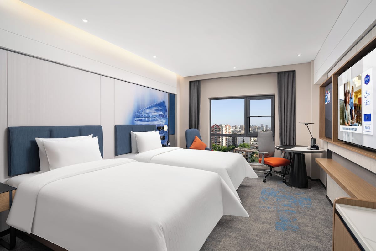 Une chambre d’hôtel avec deux lits, un bureau, une chaise, une télévision et une grande fenêtre donnant sur la ville.