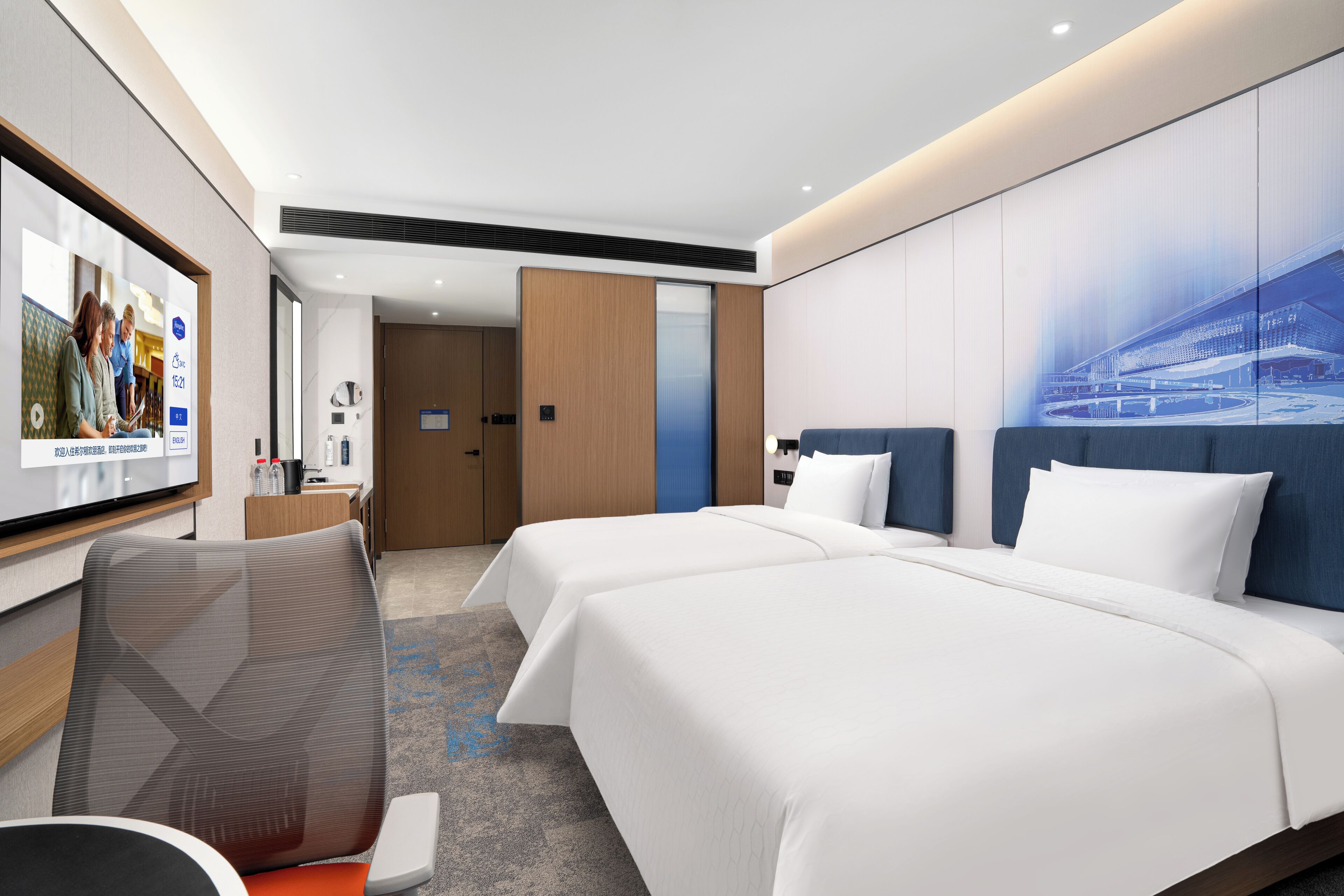 A hotel room with two beds, a desk, a chair, a large window, and a wall-mounted TV.