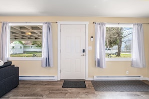A room with a door, two windows with curtains, a gray sofa, a black doormat, and a wooden floor. - New! Cherry Orchard Cottage | Fireplace · Near Flathead Lake · 1 Hr to Glacier National Park (Bigfork)