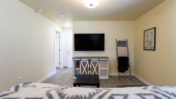 Quarto com cama, televisão presa na parede, uma mesa pequena e porta para outro espaço.
