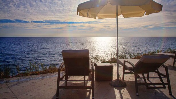 Une terrasse en bord de mer avec deux chaises longues et une table, un parasol et une vue sur l’océan.
