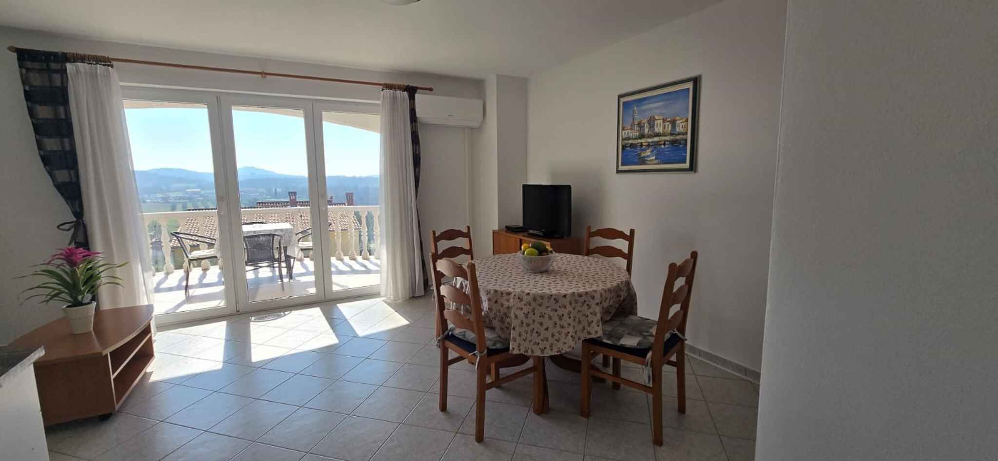 Una habitación con balcón, una mesa de comedor con sillas, un televisor y un cuadro en la pared.