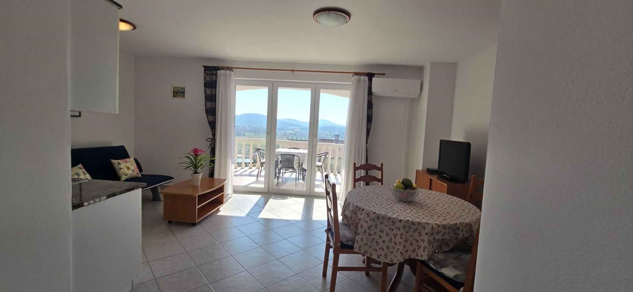 A living room with a sofa, a TV, a dining table with chairs, and a balcony with a view.
