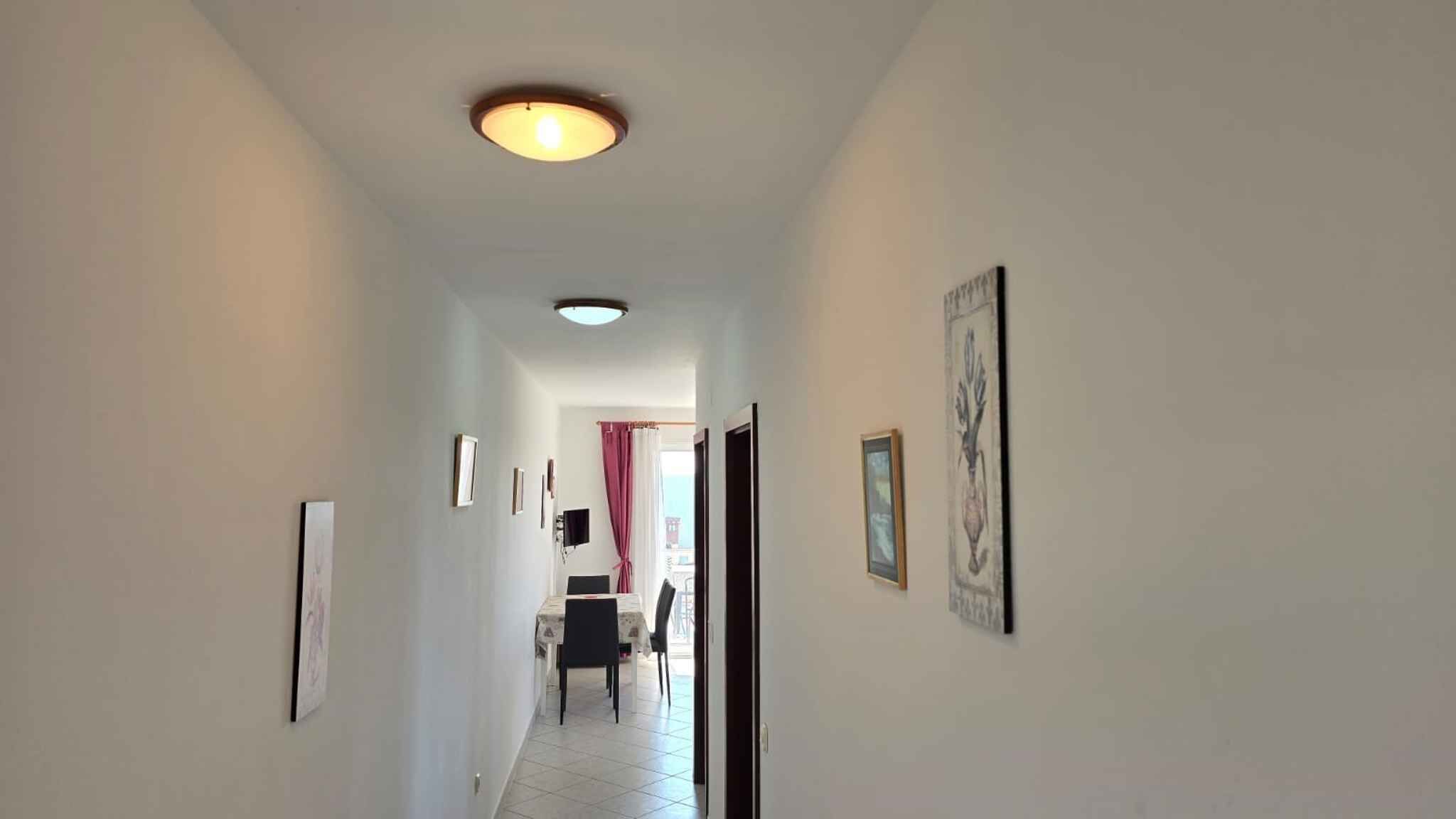 Un corridoio con una luce a soffitto, quadri alle pareti e una zona pranzo in lontananza.