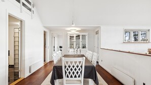 Et spiseområde med et langt bord, hvide stole, en lysekrone og et skab med glasdøre.
