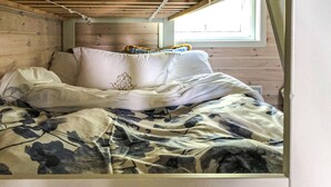 Cama com manta de camuflagem, travesseiros brancos e cabeceira de madeira em um quarto com janela.