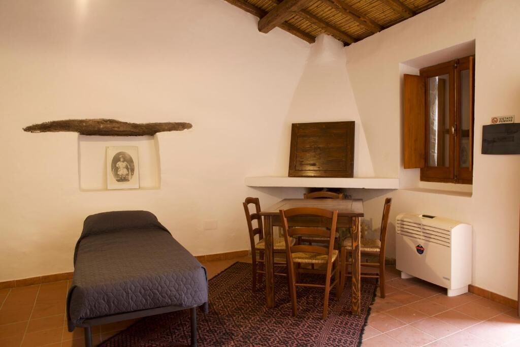 Une pièce comprenant un lit, une table avec des chaises, un radiateur et une étagère fixée au mur, ornée d’un tableau encadré et d’une plaque.