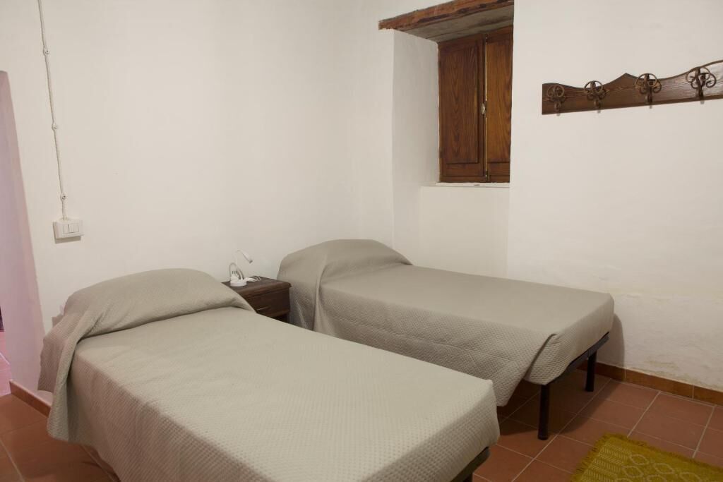Um quarto pequeno com duas camas de solteiro, um armário de madeira, uma mesinha e uma janela com moldura de madeira.