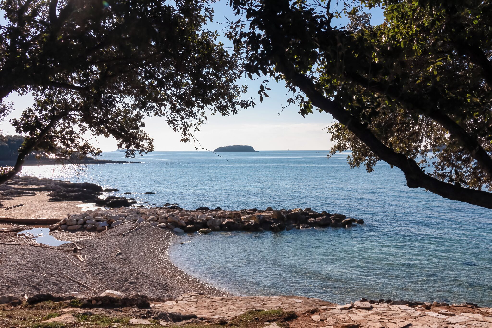 Uma praia com costa rochosa, águas calmas e uma ilha ao longe.