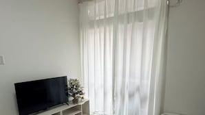 Una sala de estar con un televisor de pantalla plana sobre un mueble bajo, un sofá gris y una cortina blanca.