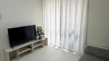 A living room with a flat-screen TV on a stand, a grey sofa, and a white curtain.