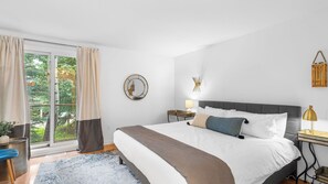 A bedroom with a large bed, a bedside table with a lamp, a round mirror, and a view of greenery outside. - Maxwell by Avantstay 15min to Village + Slopes (Vail)