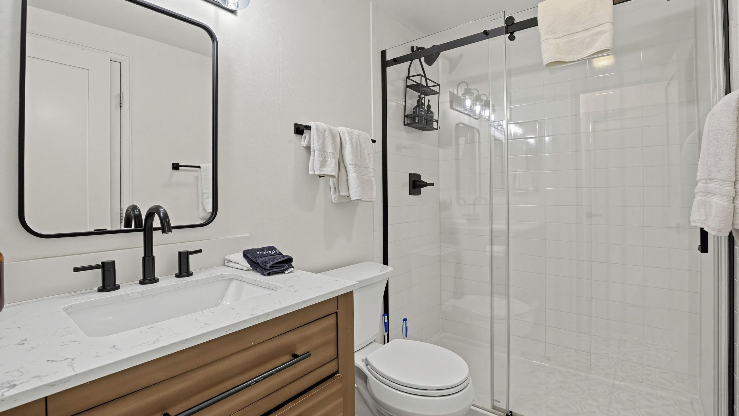 Une salle de bain moderne avec un lavabo blanc, des robinets noirs, un meuble-lavabo en bois, des toilettes et une douche en verre.