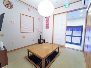 A room with tatami flooring, a wooden table, a wall clock, a window with blinds, and a sliding door. - Traditional House West Ikebukuro (Tokyo)