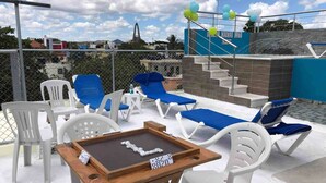 Terraço no telhado com cadeiras de descanso, uma mesa com dominós e vista para um edifício com um arco característico.