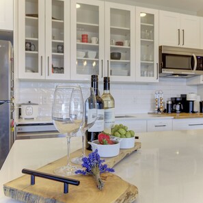 Una cocina con armarios blancos, microondas e isla de madera con copas de vino, botellas y un bol de uvas.