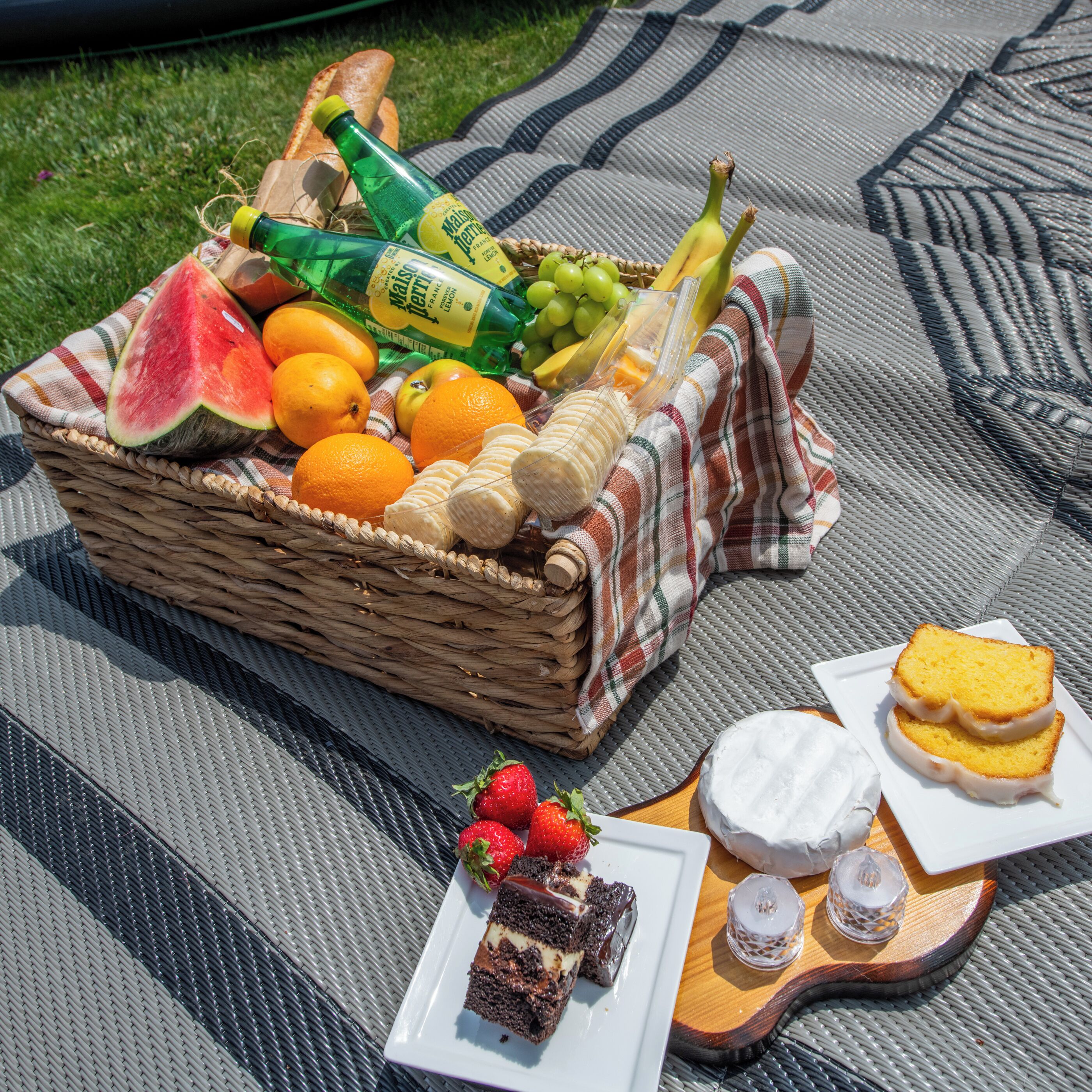 A picnic setup with a basket of fruits, bread, and a bottle of drink on a checkered blanket.