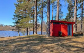 A red cabin with a small window and a door, surrounded by tall trees and a body of water.