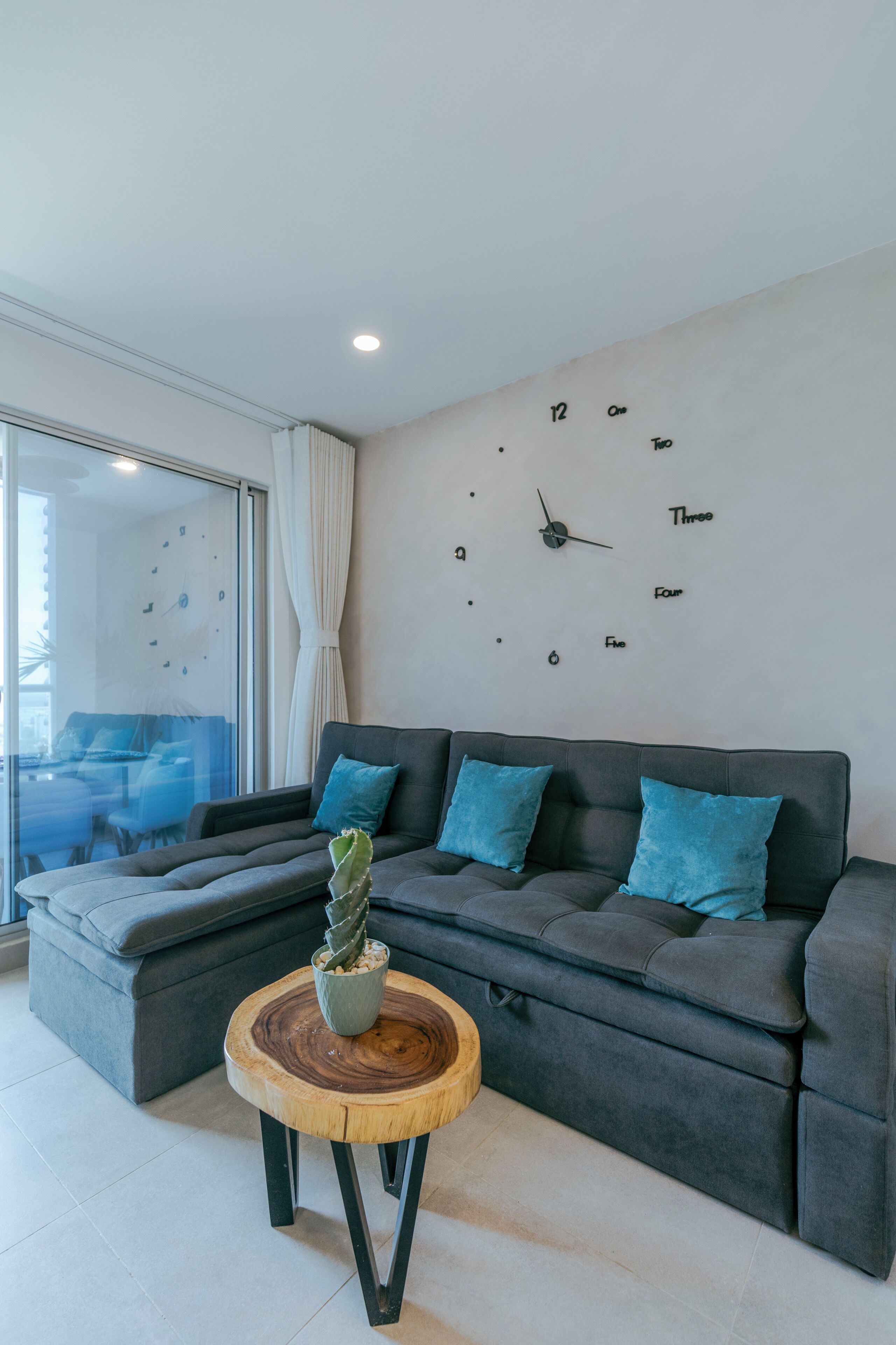 A modern living room with a grey sofa, blue cushions, a wooden coffee table with a cactus, and a wall clock.