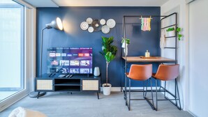 A modern living room with a flat-screen TV, a dining area with two chairs, a small table, a potted plant, and a wall-mounted shelf with decorative items.