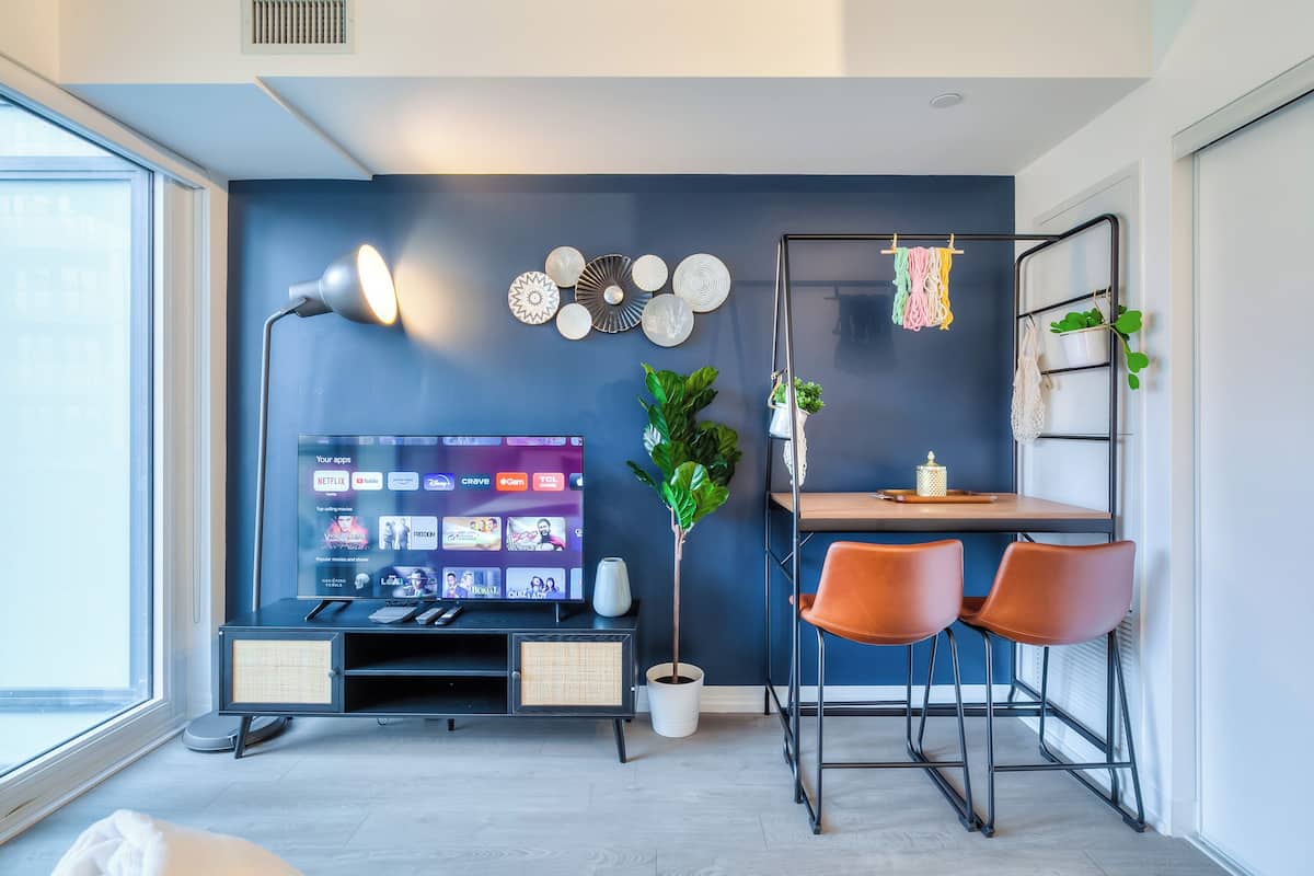 Una sala moderna con televisor de pantalla plana, un área de comedor con dos sillas, una mesita, una planta en maceta y un estante en la pared con objetos decorativos.